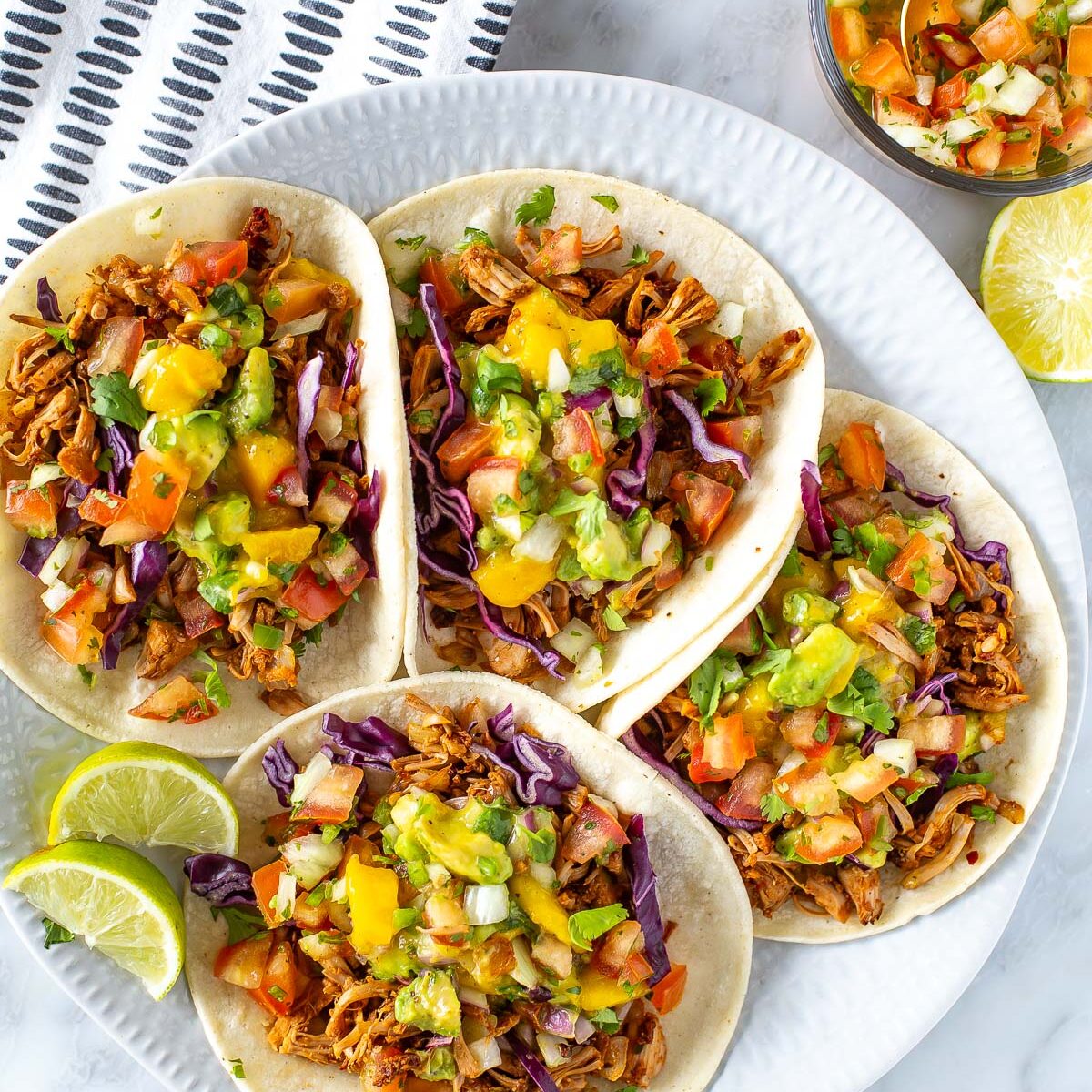 Overhead shot of four jackfruit tacos on a white plate, topped with jackfruit, mango avocado salsa, and red cabbage. Lime wedges and a small bowl of cilantro are also visible on a marble surface.