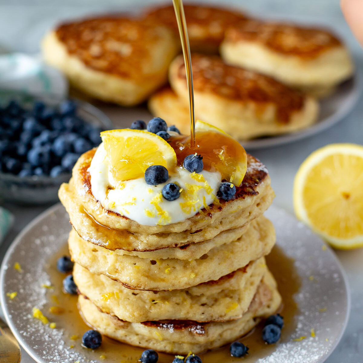 Side shot of a tall stack of Lemon Ricotta Pancakes being drizzled with maple syrup from a glass bottle.