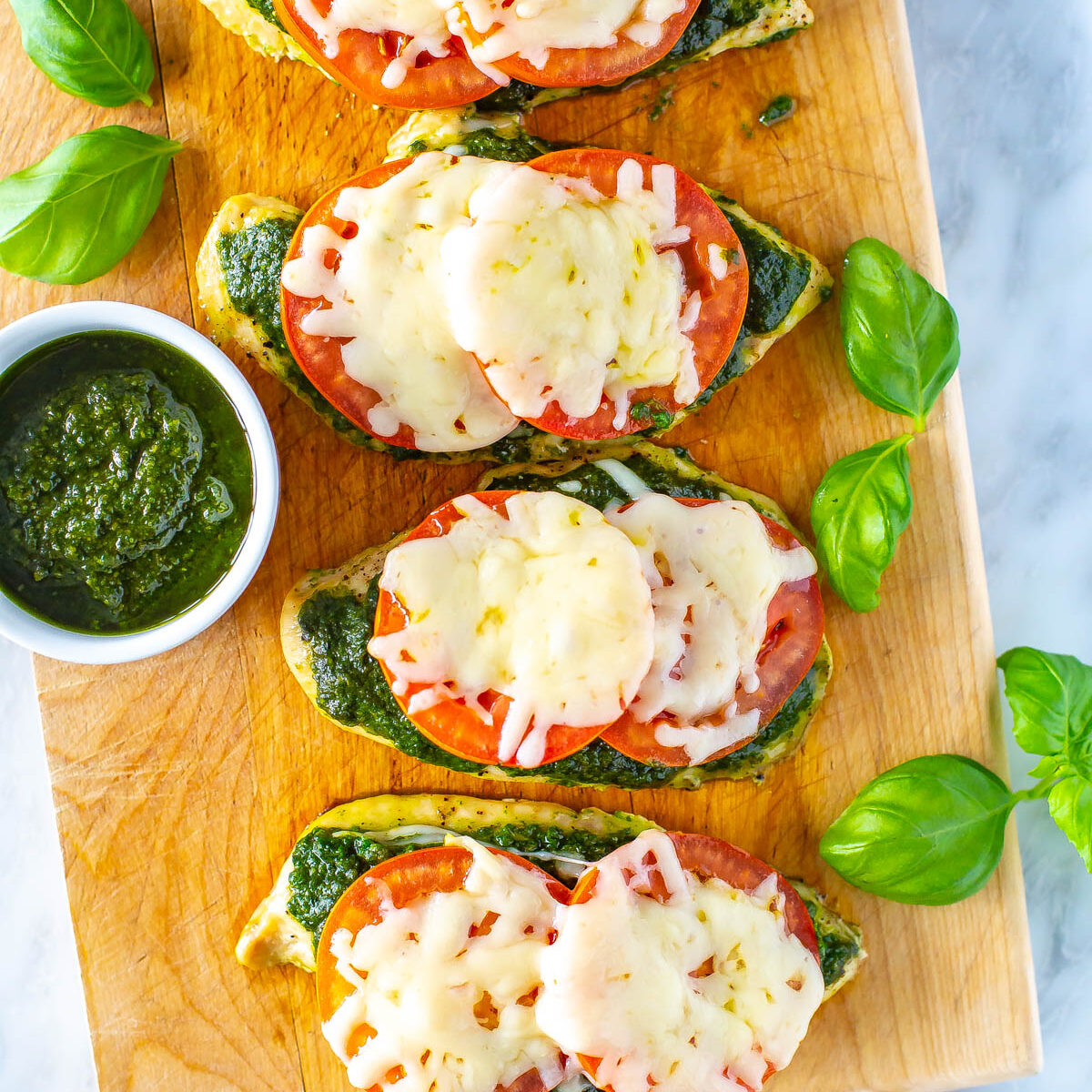 Four chicken cutlets on a butcher block topped with pesto, tomato and mozzarella cheese.