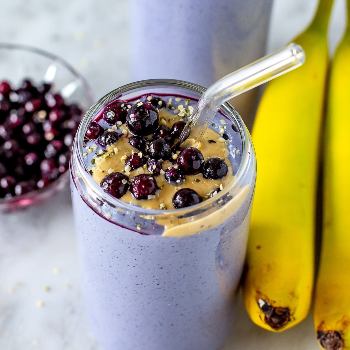 Overhead shot of two purple tofu smoothies topped with blueberries and peanut butter, with bananas on the side.