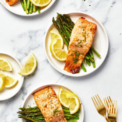A zoomed-out shot with three plates, each with a bed of asparagus topped with air fryer salmon, garnished with lemon slices.